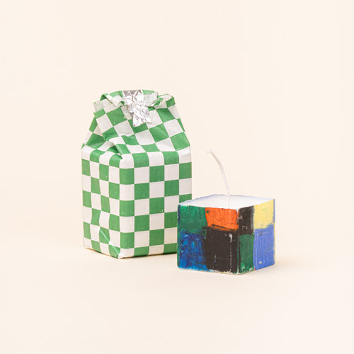 A short, square hand-painted candle shown beside its green-and-white checkerboard paper packaging. The candle features bold blocks of green, blue, black, orange, and yellow, with a white wax top and long wick.