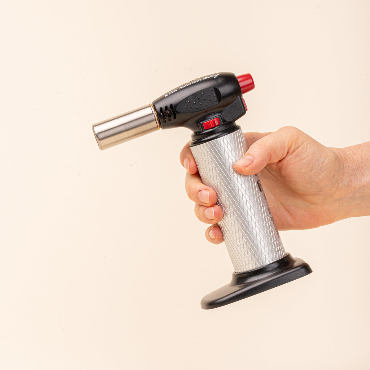 A hand holds a black and silver culinary torch with red controls on a light background.