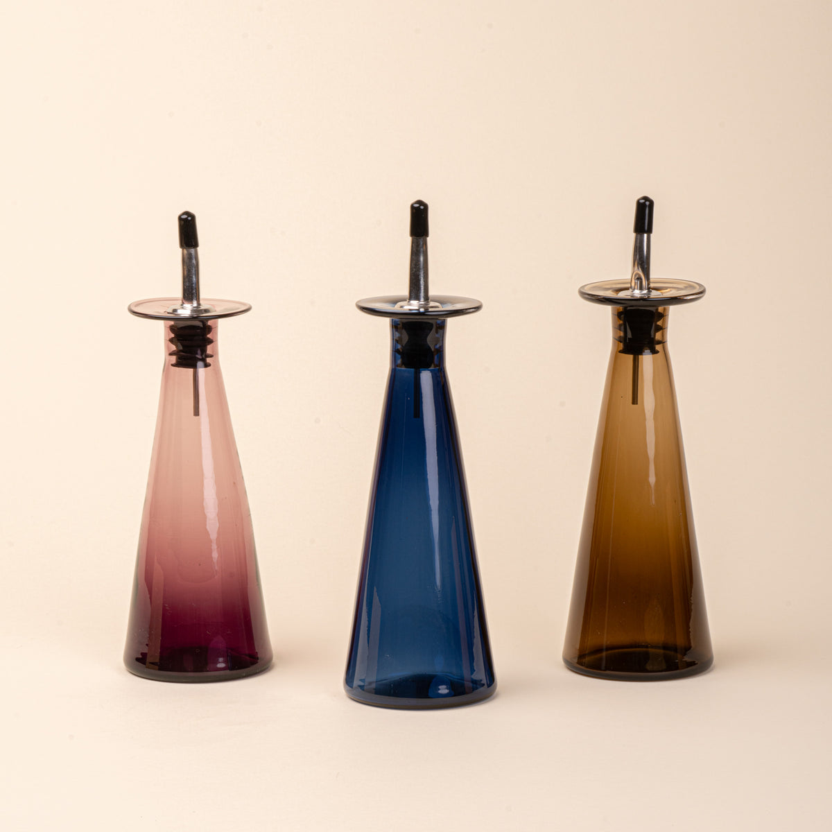 Three glass oil or vinegar dispensers with a tapered bottle shape, wide flared collar, and metal pour spout with a black stopper, in plum, blue and amber colors.
