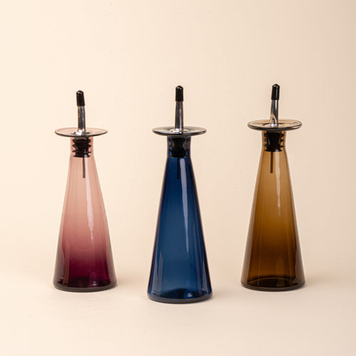 Three glass oil or vinegar dispensers with a tapered bottle shape, wide flared collar, and metal pour spout with a black stopper, in plum, blue and amber colors.