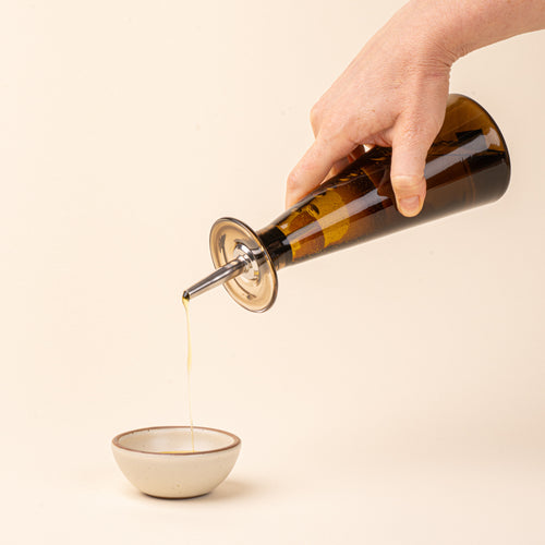 A hand holds an amber-brown glass oil or vinegar dispenser with a tapered bottle shape, wide flared collar, and metal pour spout with a black stopper, pouring oil into a small ceramic bowl.