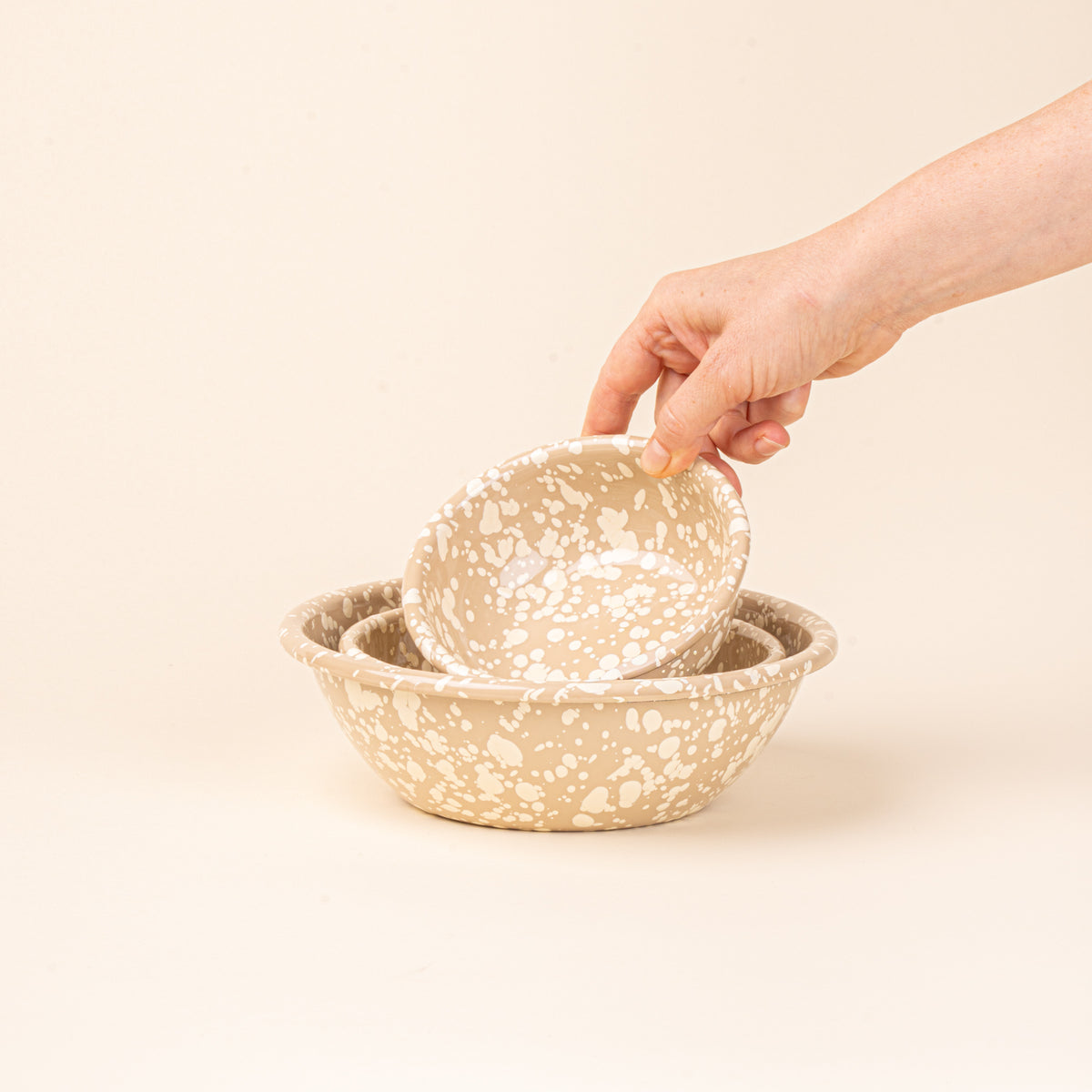 A hand holds a small enamel bowl to nest with a medium and large bowl in neutral tan color with off-white splatter pattern.