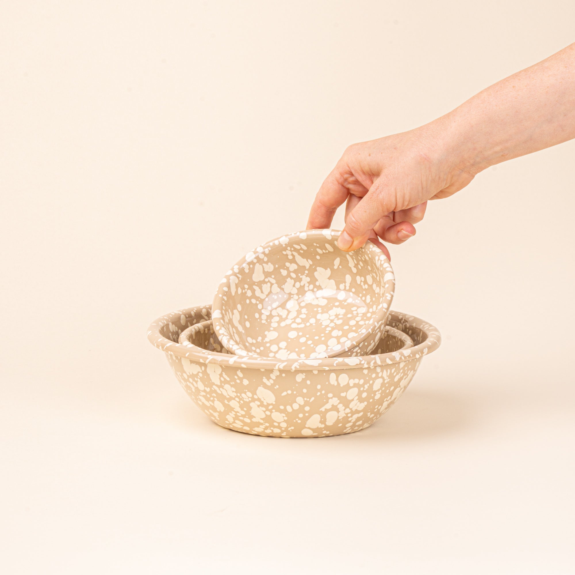 A hand holds a small enamel bowl to nest with a medium and large bowl in neutral tan color with off-white splatter pattern.