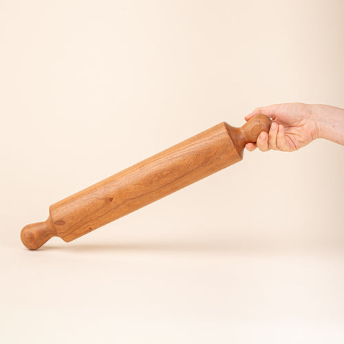 A hand holds a simple dowel wooden rolling pin with rounded handles, in cherry.