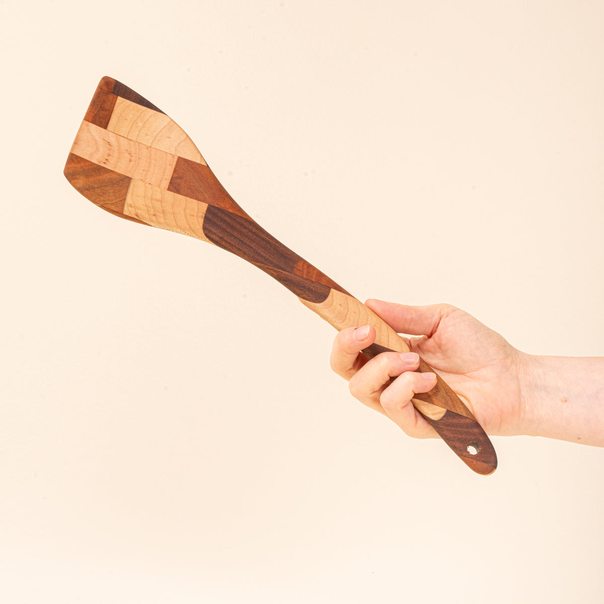 A hand holds a mixed wood spatula with a slanted head and a hole at the bottom 