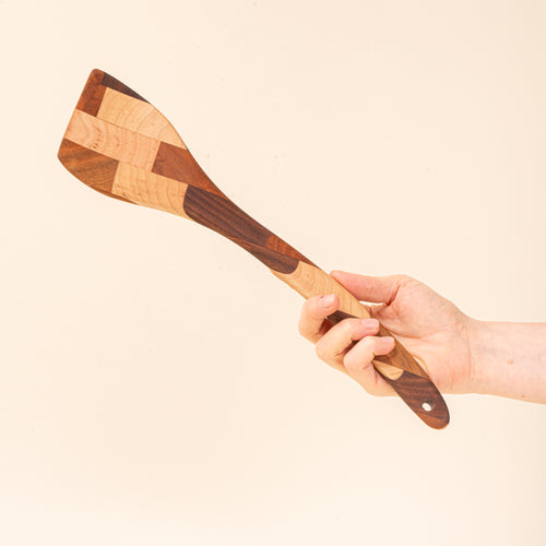 A hand holds a mixed wood spatula with a slanted head and a hole at the bottom 