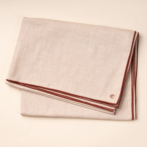 A folded beige linen tablecloth with terracotta-colored piping and embroidered monogram.