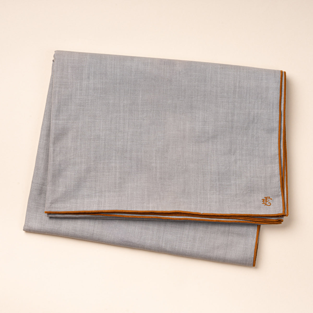 A folded light blue linen tablecloth with rust-colored piping and embroidered monogram.