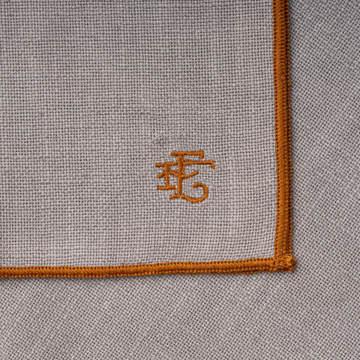 Corner of a folded light blue table linen with rust-colored piping and embroidered monogram.
