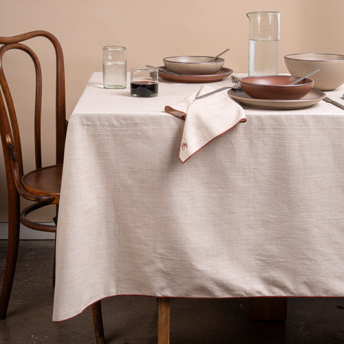 A table set with ceramic pottery, glasses of wine and water, silverware, and a beige table linen with terracotta-colored piping and embroidered monogram.
