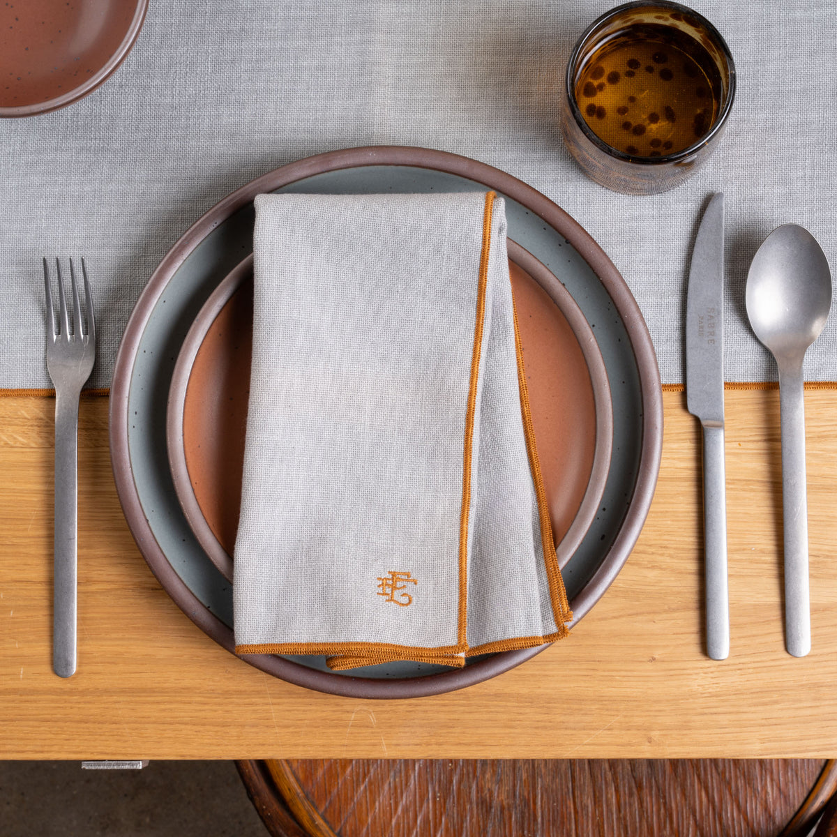 A place setting with a folded light blue table napkin with rust-colored piping and embroidered monogram, with plates in a complementary colorway.