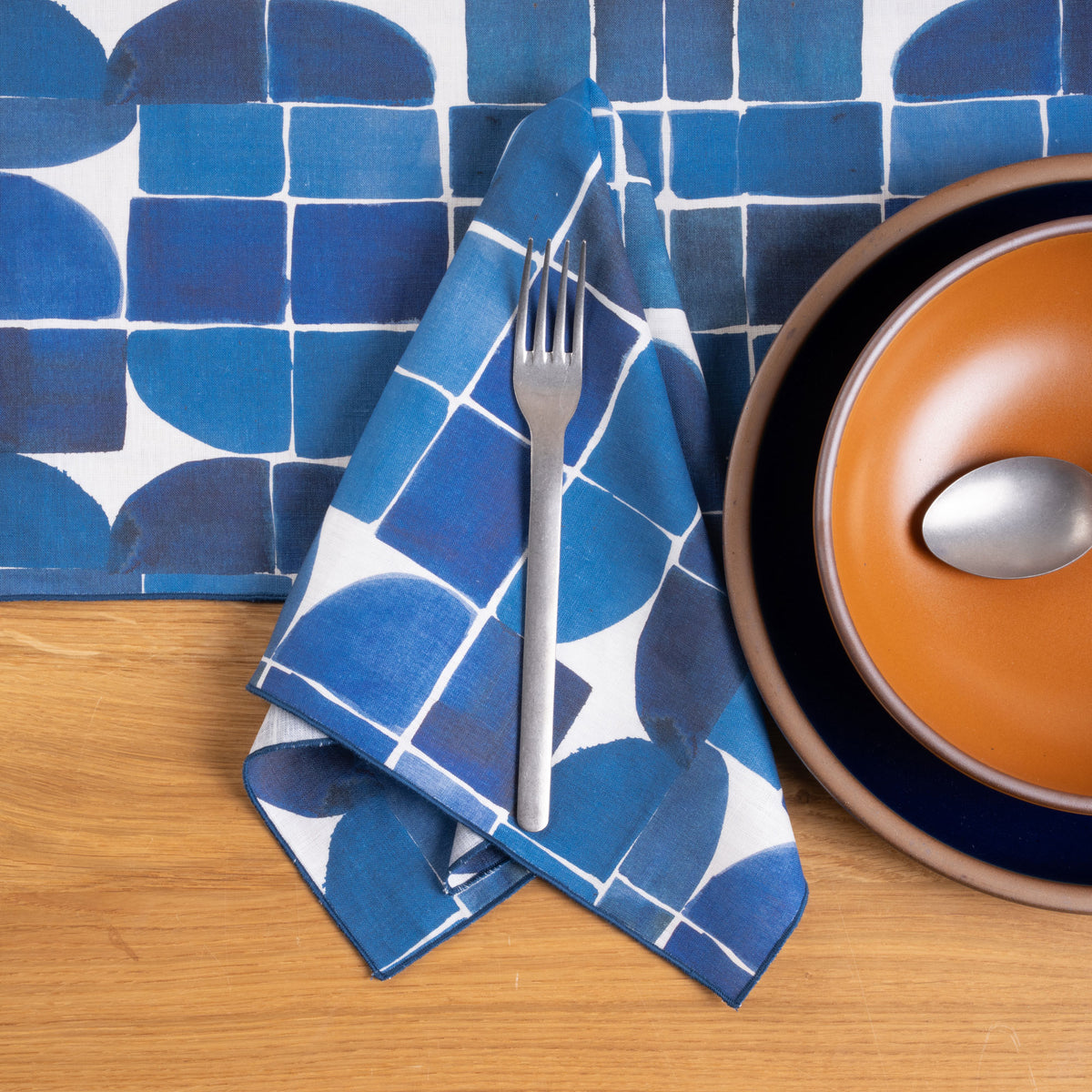 Blue geometric napkin and tablecloth paired with warm orange and deep navy dinnerware on a wood table.