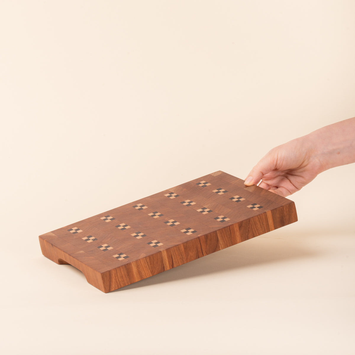 A hand holds out a rectangular wooden cutting board with repeating geometric inlay pattern on a light background.