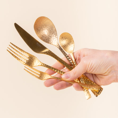 A hand holds a hammered stainless steel 5 piece flatware in a gold color