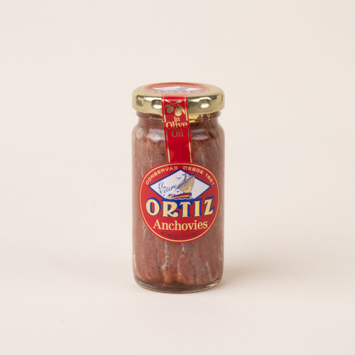 Glass jar of Ortiz anchovies in olive oil with a gold lid and red, blue, and gold label on a cream background.