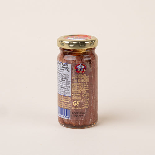 Back view of a glass jar of Ortiz anchovies in olive oil with a gold lid, showing the nutrition facts and product label.