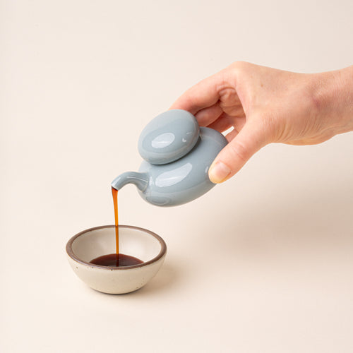 A hand holds a pale blue glazed ceramic soy sauce pot with a low, rounded body, short pouring spout, and domed lid, pouring soy sauce into a small bowl.