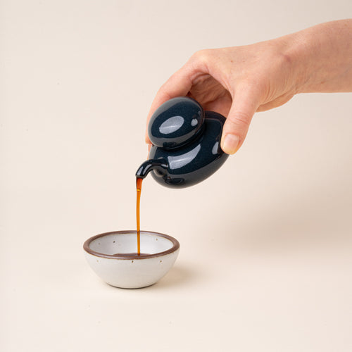 A hand holds a navy glazed ceramic soy sauce pot with a low, rounded body, short pouring spout, and domed lid, pouring soy sauce into a small bowl.
