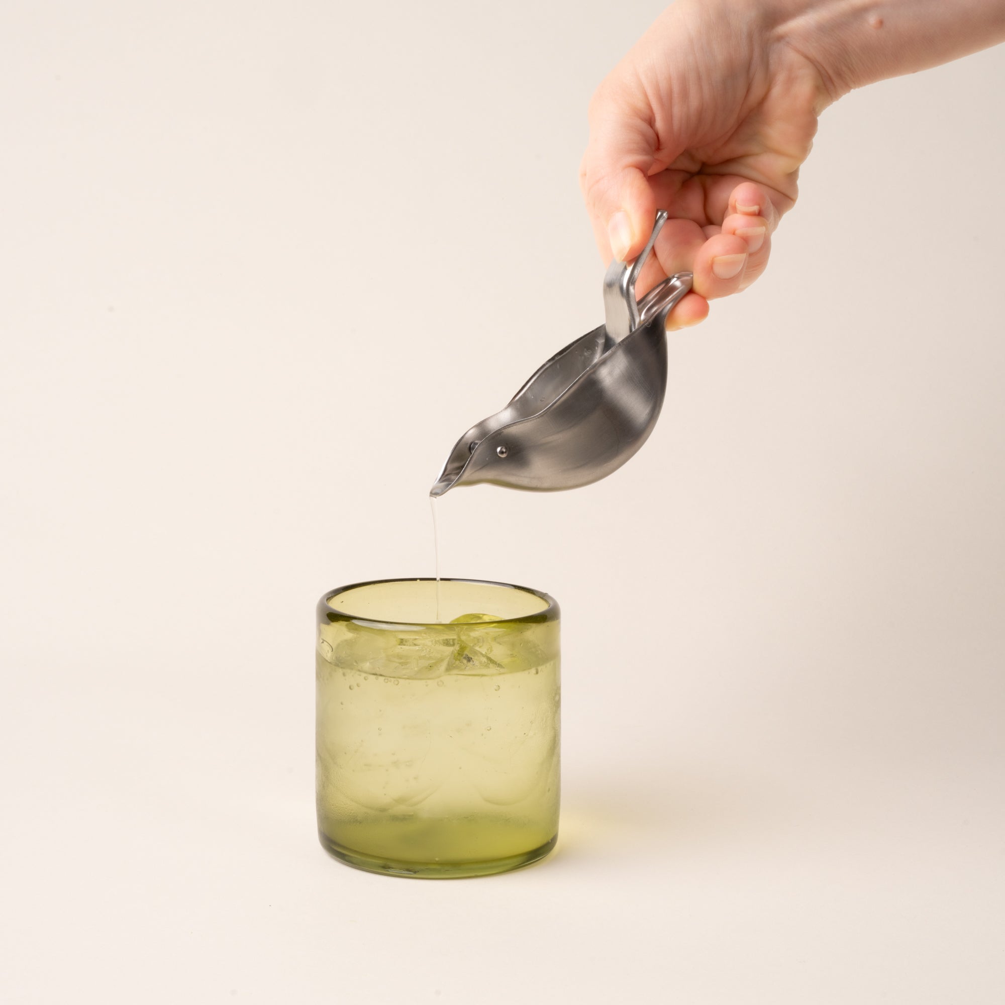 A hand holds a bird-like silhouetted lemon squeezer pouring juice into a glass of water.