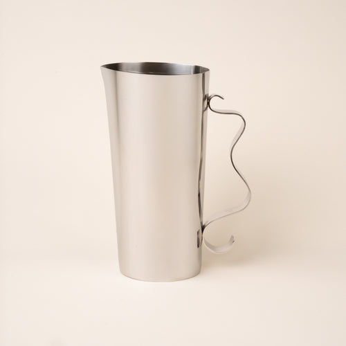A polished steel pitcher with a tall cylindrical body, subtle pouring spout, and sculptural curved handle, photographed against a warm neutral background.