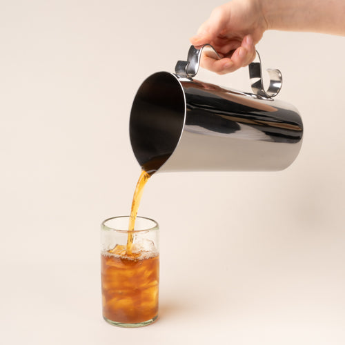 A polished steel pitcher held by its sculptural handle, pouring amber iced tea into a clear glass against a warm neutral background.