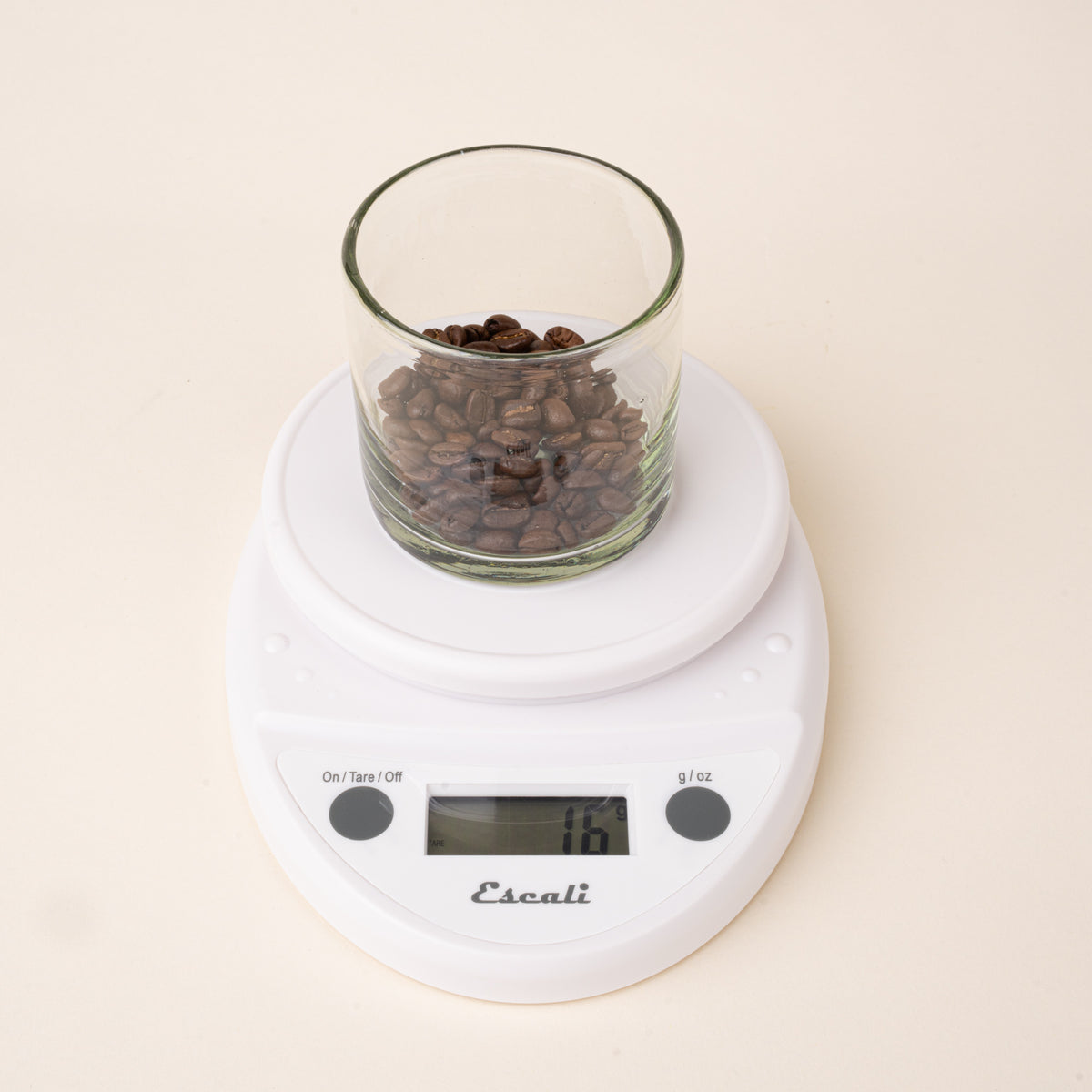 Coffee beans in a glass on a white digital kitchen scale with a round weighing platform and LCD display.