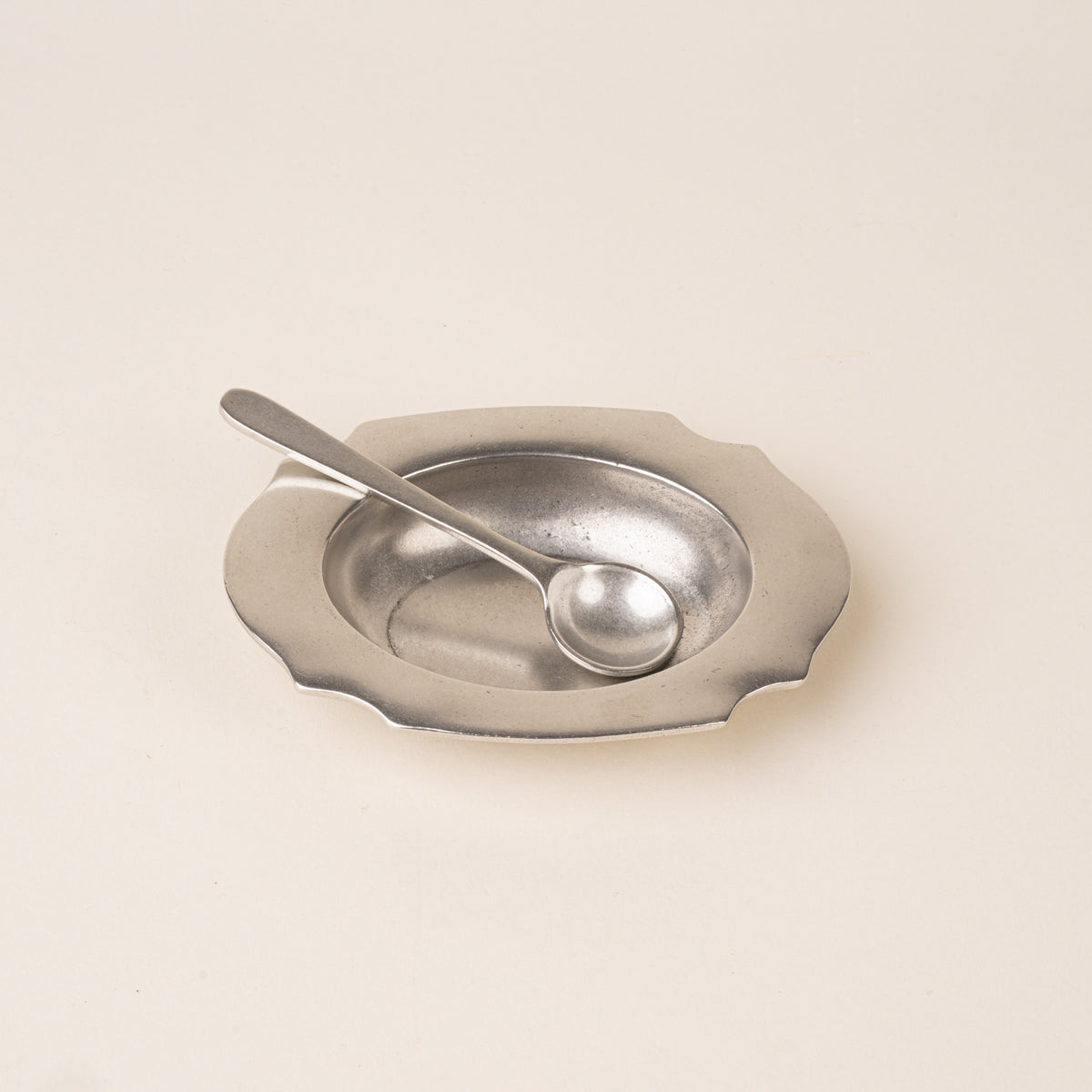 A pewter salt cellar with a broad scalloped rim, paired with a small matching serving spoon.