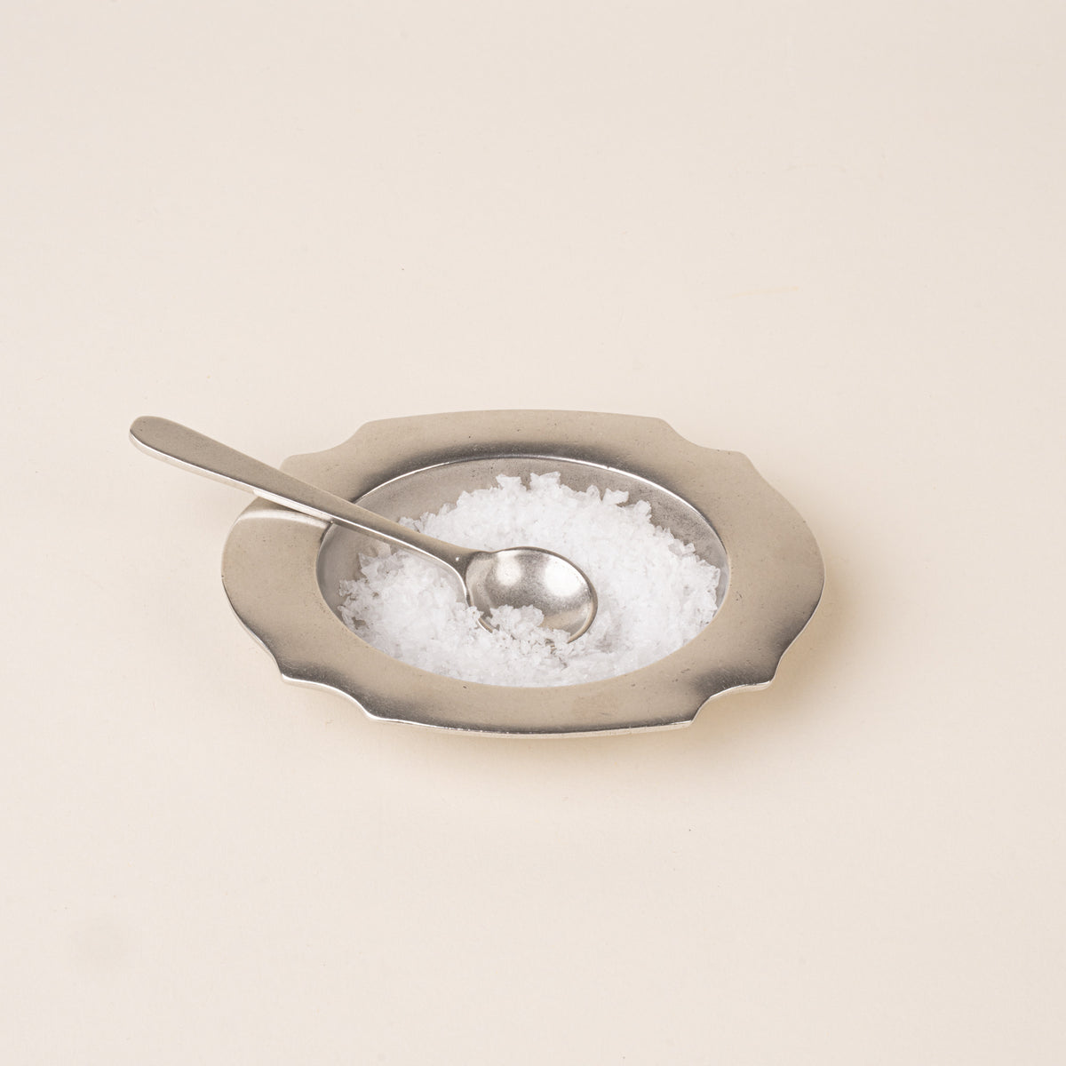A pewter salt cellar with a broad scalloped rim, filled with salt and paired with a small matching serving spoon.
