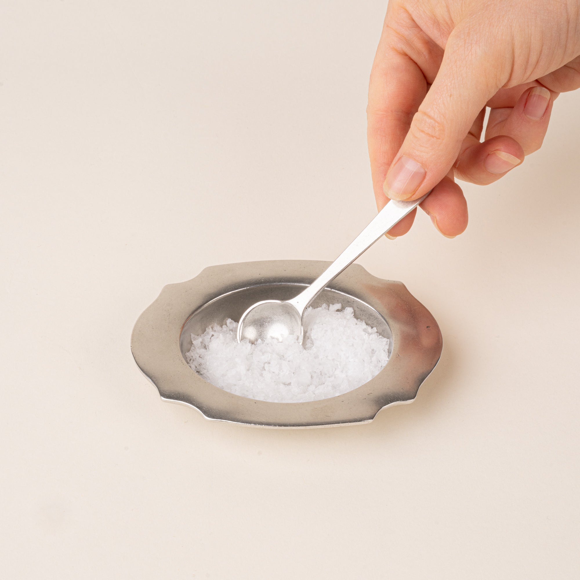 A pewter salt cellar with a broad scalloped rim, filled with salt and a hand holding a small matching serving spoon.