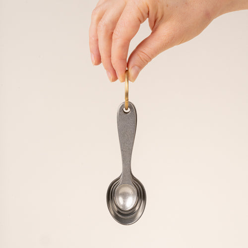 A hand holds four pewter measuring spoons on a brass ring nested together.