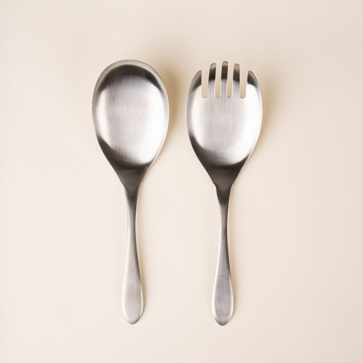 A stainless-steel salad serving set with an oversized spoon and four-tined serving fork, featuring smooth oval heads and gently tapered handles, photographed on a warm cream background.