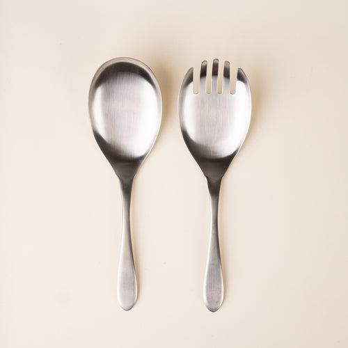 A stainless-steel salad serving set with an oversized spoon and four-tined serving fork, featuring smooth oval heads and gently tapered handles, photographed on a warm cream background.