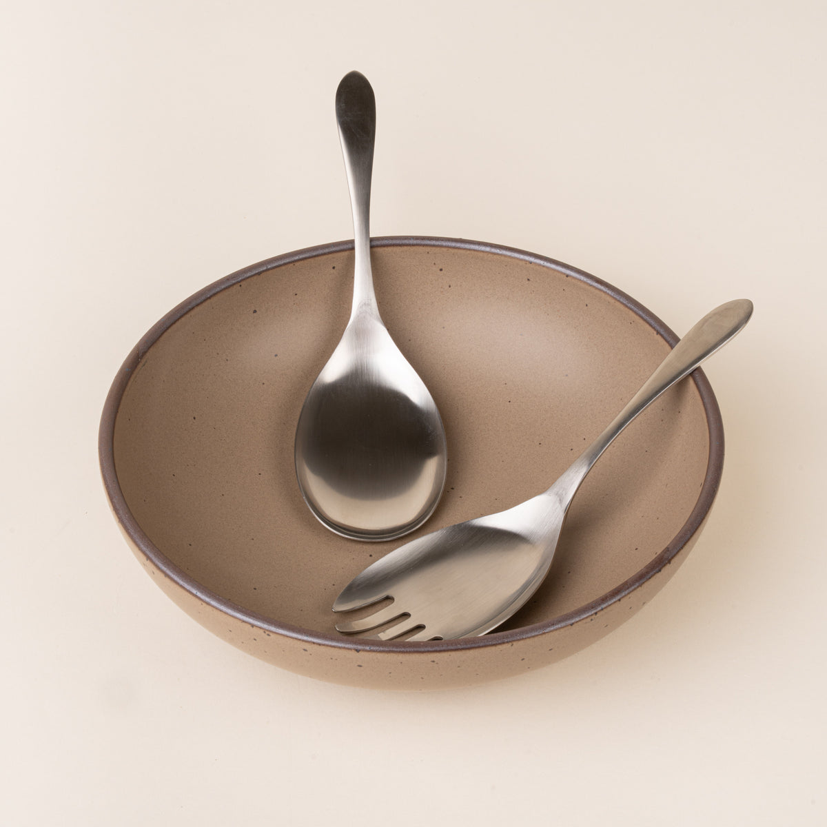 A stainless-steel salad serving set with an oversized spoon and four-tined serving fork, featuring smooth oval heads and gently tapered handles sitting in a large serving board.
