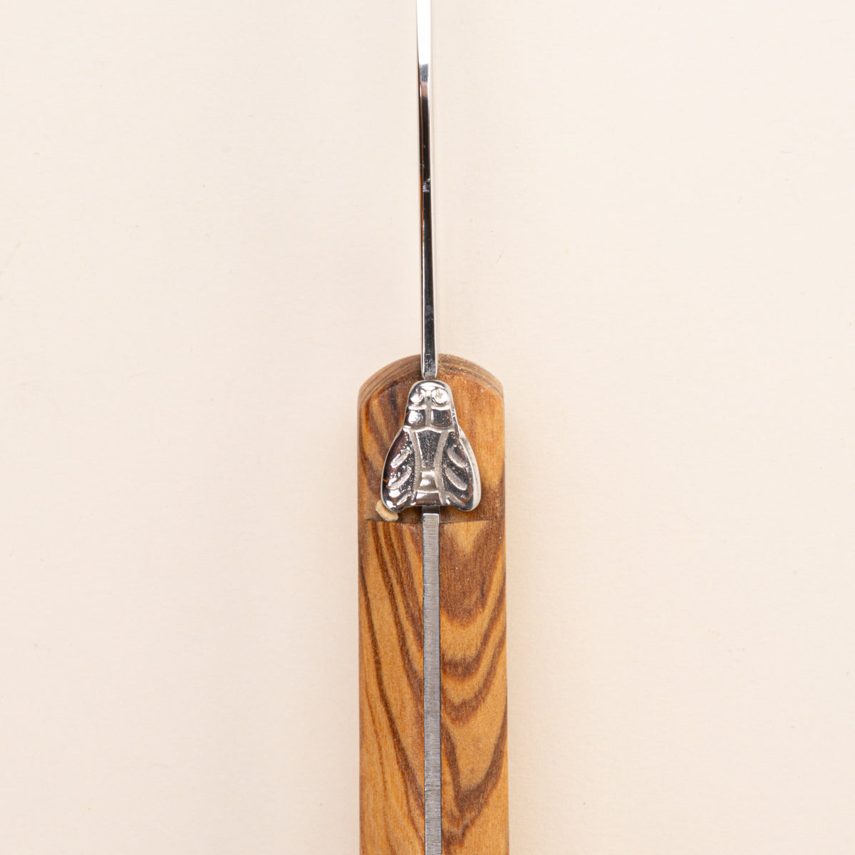 A close-up of a knife handle made of warm-toned olive wood with a polished silver metal blade and a decorative silver bee emblem on the guard.