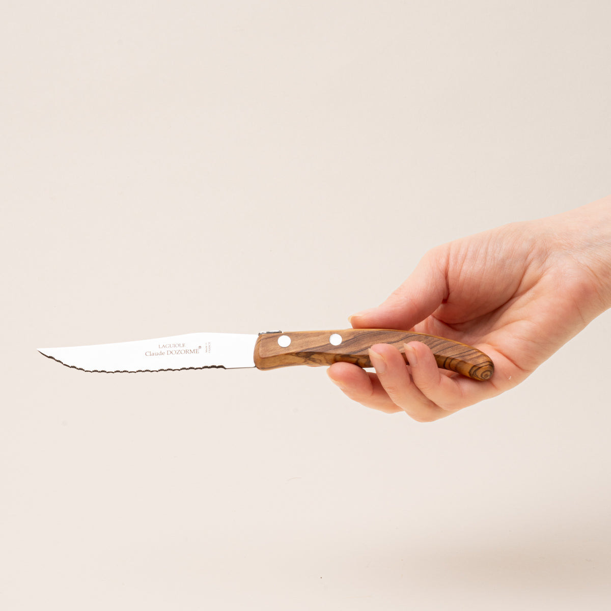 A hand holds a steak knife with polished steel blades and richly grained olive wood handle.