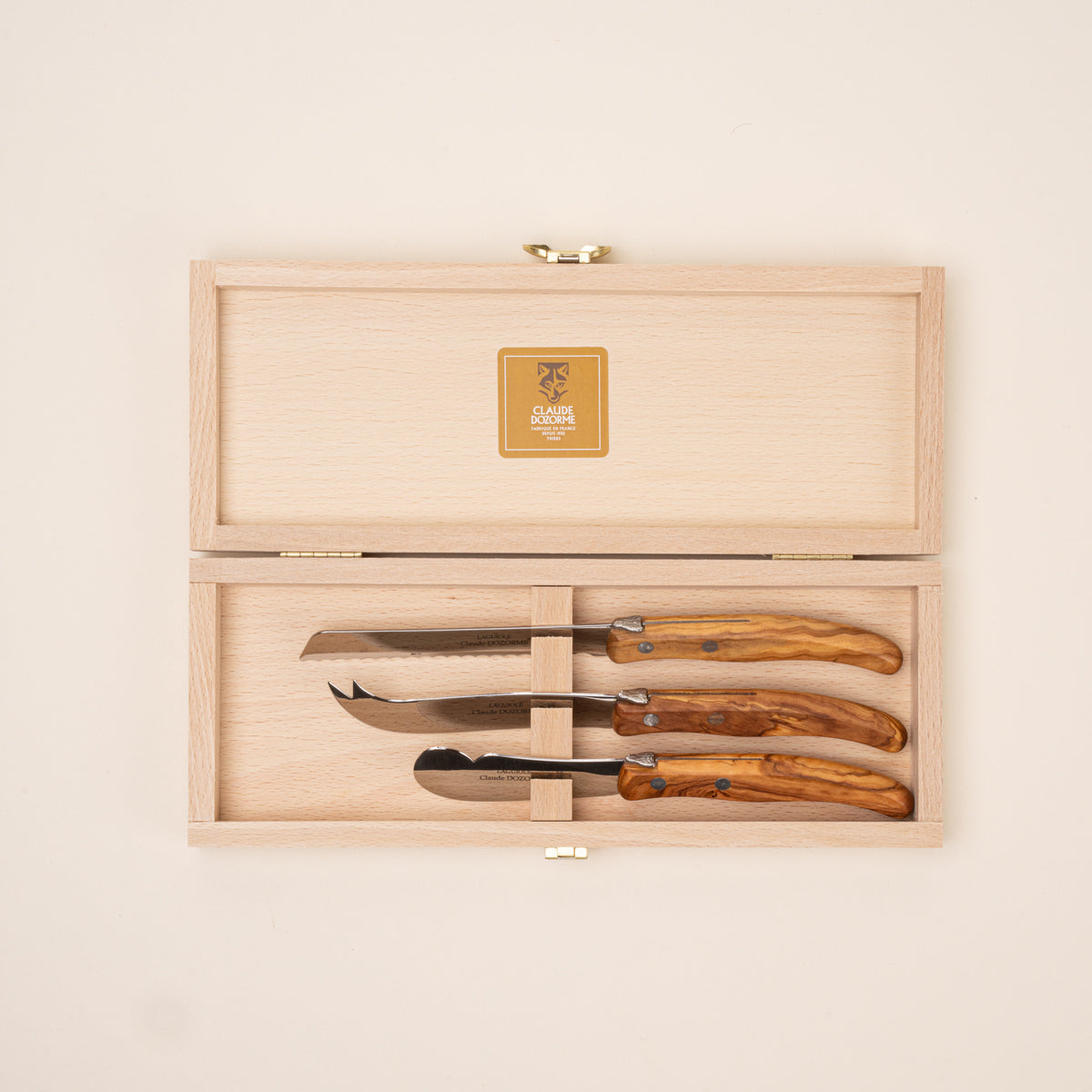Set of three serving knives with polished steel blades and olive wood handles, including cheese, bread, and spreader knives, sitting in a wooden open box.