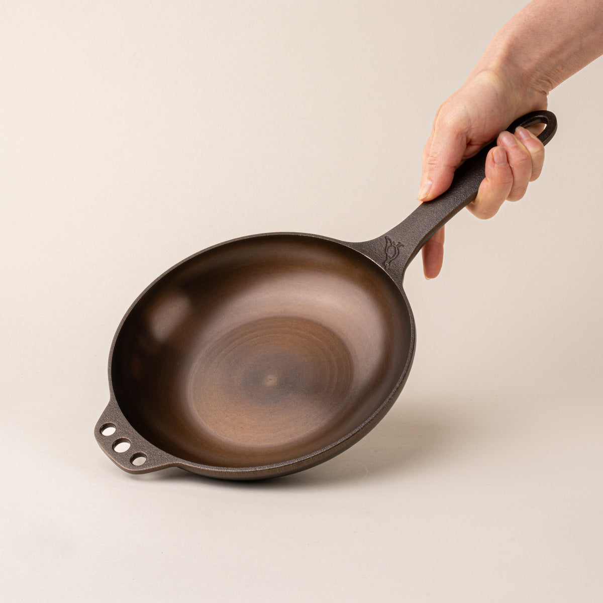 Hand holding the handle of a dark brown small cast iron skillet with a smooth cooking surface and three holes on the opposite side.