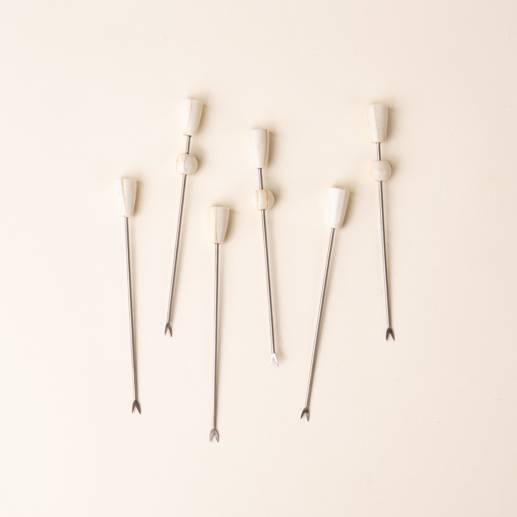 Six slender metal cocktail or appetizer picks with small two-pronged fork tips and sculptural ivory-colored handles, arranged vertically on a warm cream background.
