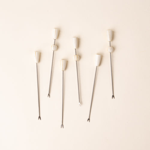 Six slender metal cocktail or appetizer picks with small two-pronged fork tips and sculptural ivory-colored handles, arranged vertically on a warm cream background.