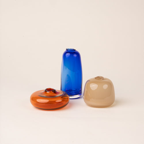 Three small handblown glass bud vases in contrasting colors and shapes: a low amber-orange form, a tall cobalt-blue bottle, and a rounded smoky-beige vessel, arranged on a warm cream background.