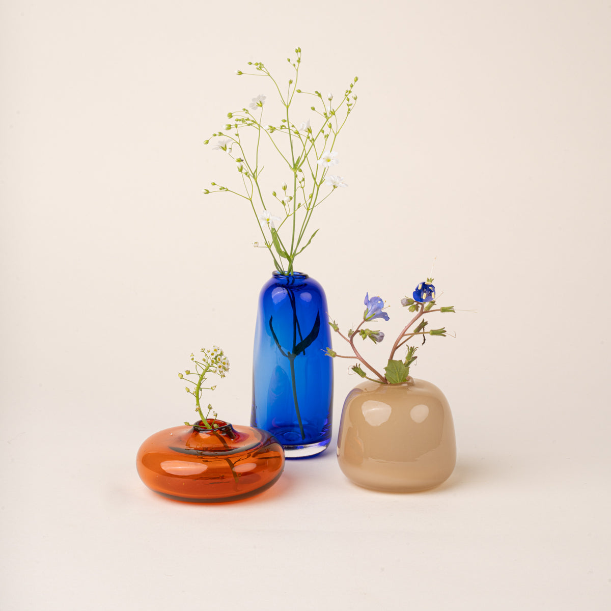 Three small handblown glass bud vases filled with small flowers, in contrasting colors and shapes: a low amber-orange form, a tall cobalt-blue bottle, and a rounded smoky-beige vessel, arranged on a warm cream background.