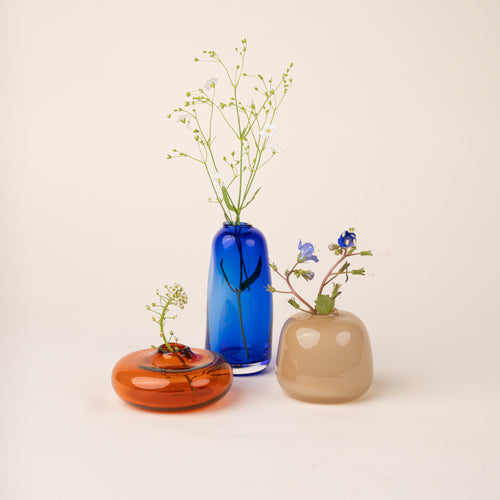 Three small handblown glass bud vases filled with small flowers, in contrasting colors and shapes: a low amber-orange form, a tall cobalt-blue bottle, and a rounded smoky-beige vessel, arranged on a warm cream background.