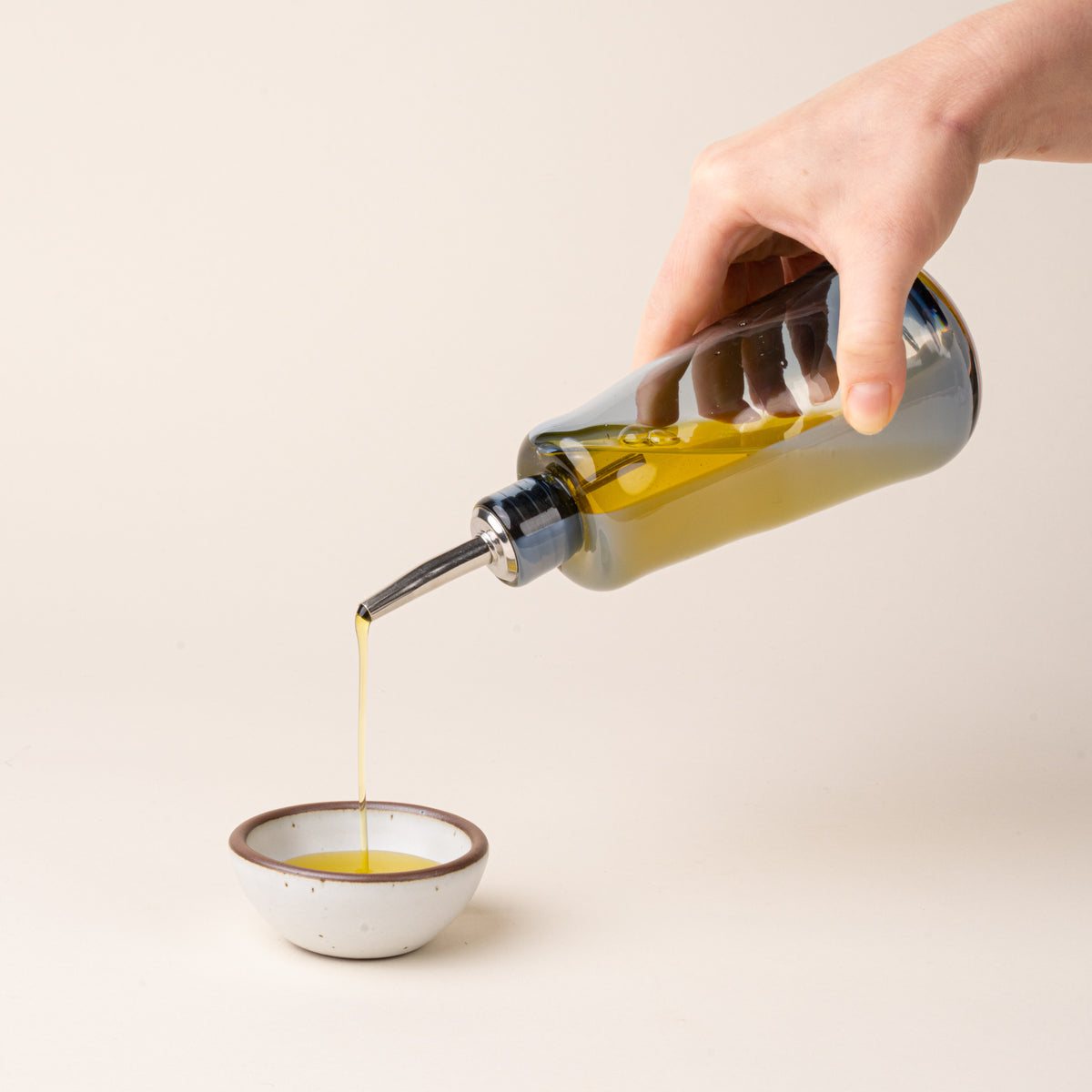 A hand holds a blue-gray glass oil or vinegar dispenser with a polished metal pour spout and stopper, pouring oil into a small white ceramic bowl.