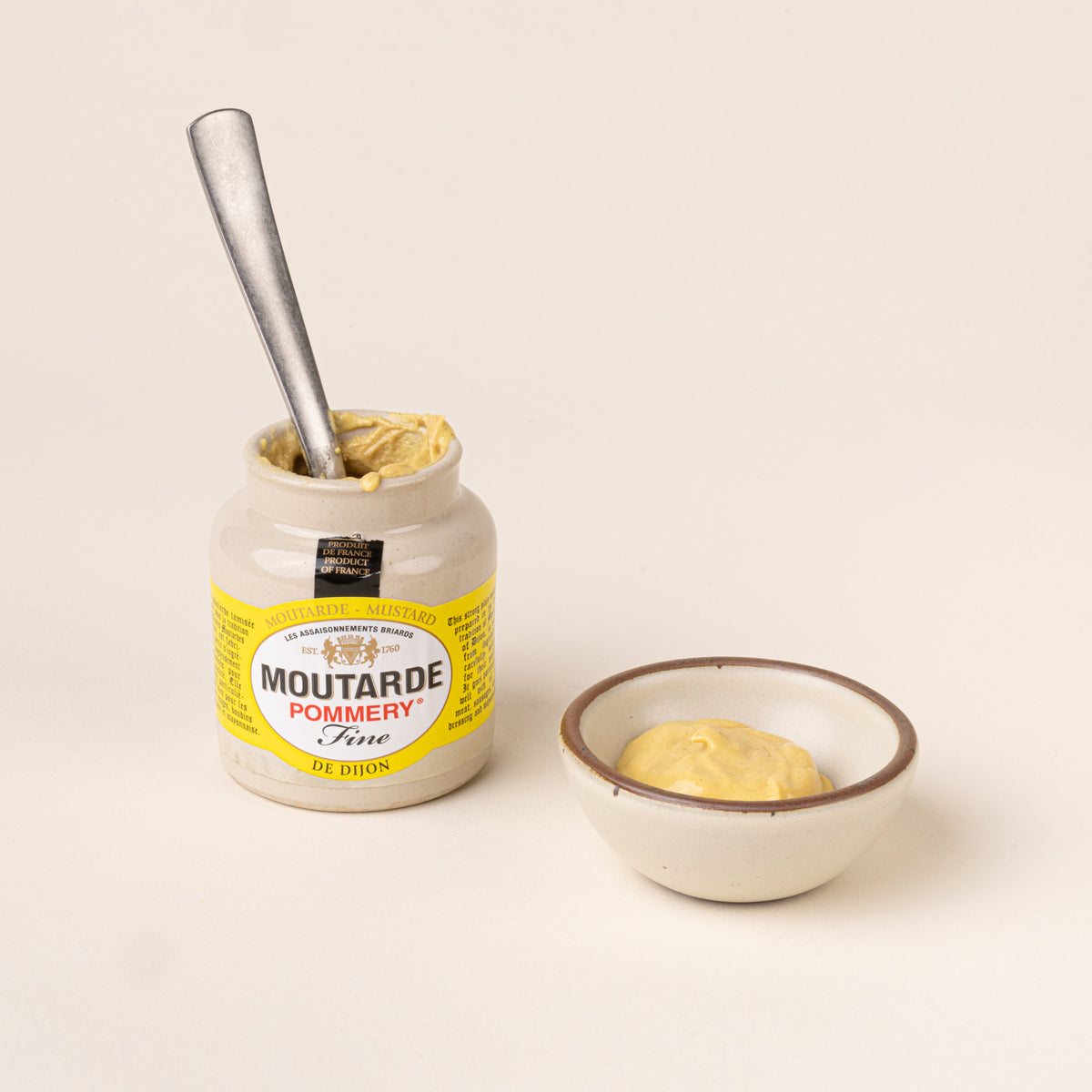 Open jar of Pommery Fine Dijon mustard with a metal spoon inside, beside a small ceramic bowl filled with mustard.