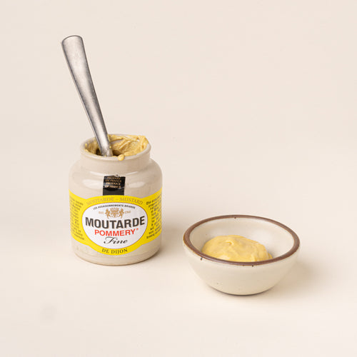 Open jar of Pommery Fine Dijon mustard with a metal spoon inside, beside a small ceramic bowl filled with mustard.