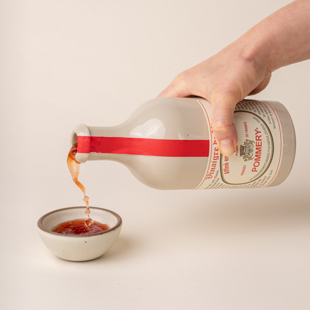 A hand pours Pommery red wine vinegar from a white ceramic bottle with a red ribbon into a small cream-colored bowl.
