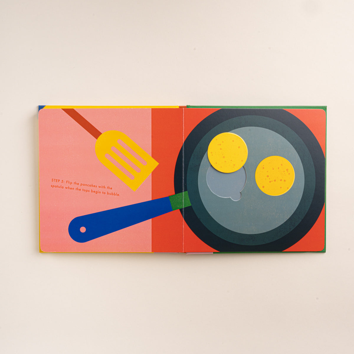 Open interactive pancake recipe book showing illustrated pancakes cooking in a pan with a spatula.