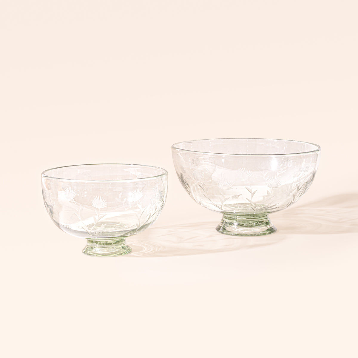 Two clear glass footed bowls in different sizes with etched floral designs bases on a cream background.