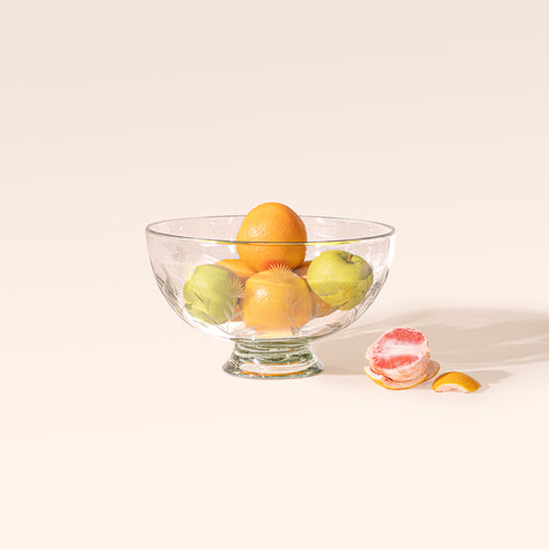 A larger clear glass footed bowl with etched floral designs, filled with grapefruits and apples on a cream background.