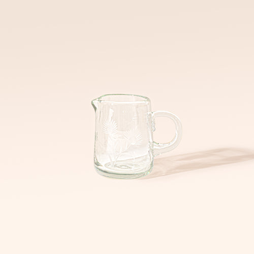 Clear glass creamer with etched floral designs, a rounded handle, and a pouring spout on a cream background.